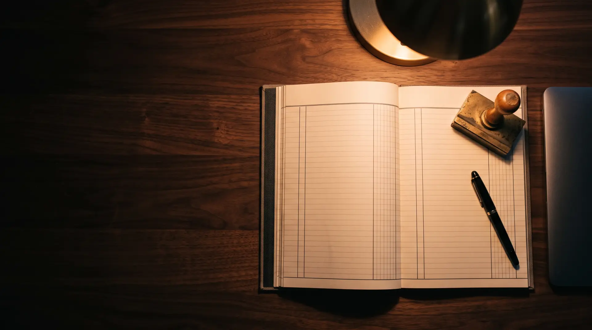 An open ledger, a pen and an ink stamp on a dark wooden desk under a lamp.